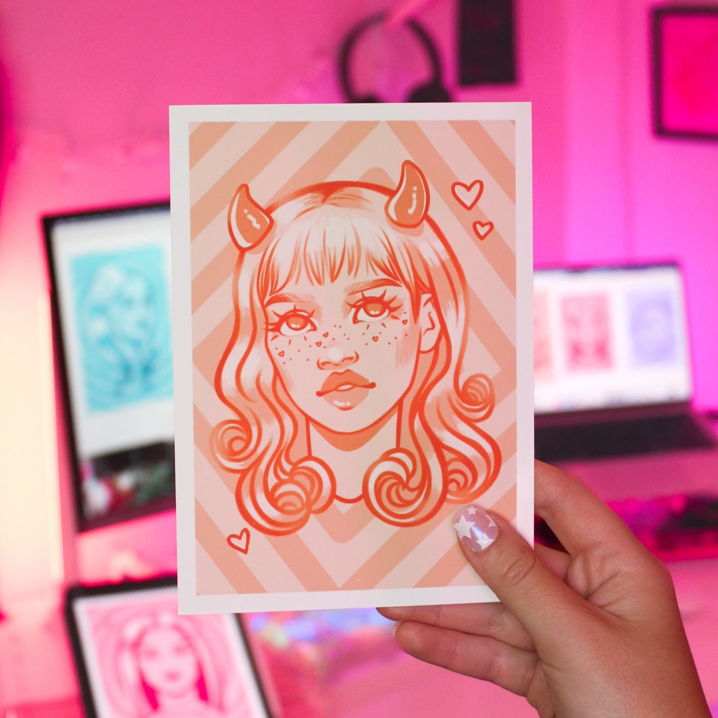 Person holding a 5x7 art print with an orange stylized illustration of a woman with horns and heart freckles in a colorful room.