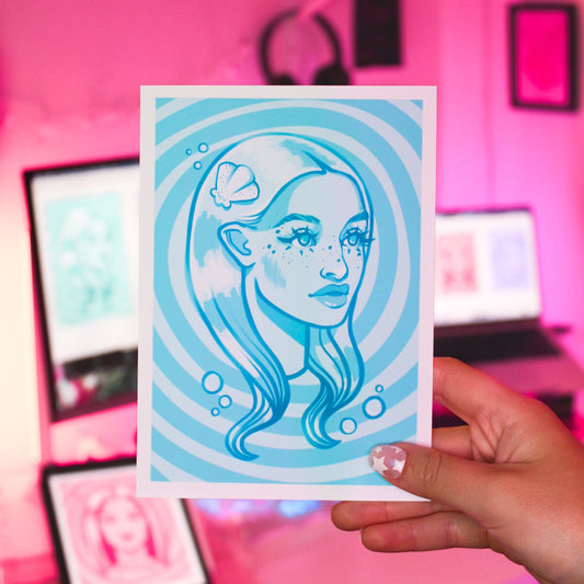 Hand holding a blue and white 5x7 art print illustration of a mermaid woman with a shell in her hair.