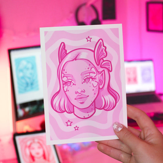 Hand holding a pink illustration of a fairy woman with fairy wings against a blurred pink-themed room background.