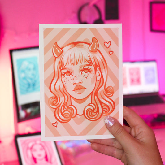 Person holding a 5x7 art print with an orange stylized illustration of a woman with horns and heart freckles in a colorful room.