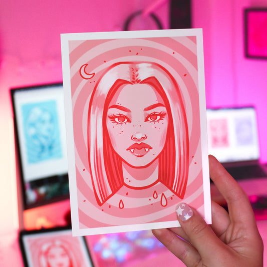 Person holding a red-themed artwork print with a stylized vampire face design against a blurred indoor background.