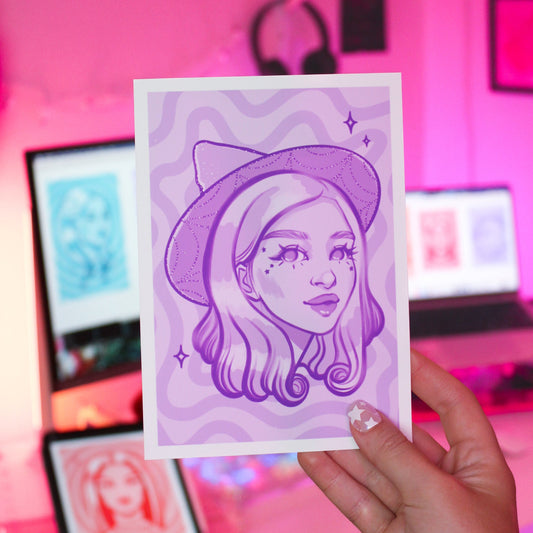 Hand holding a purple art print of a witch woman with a witch hat.
